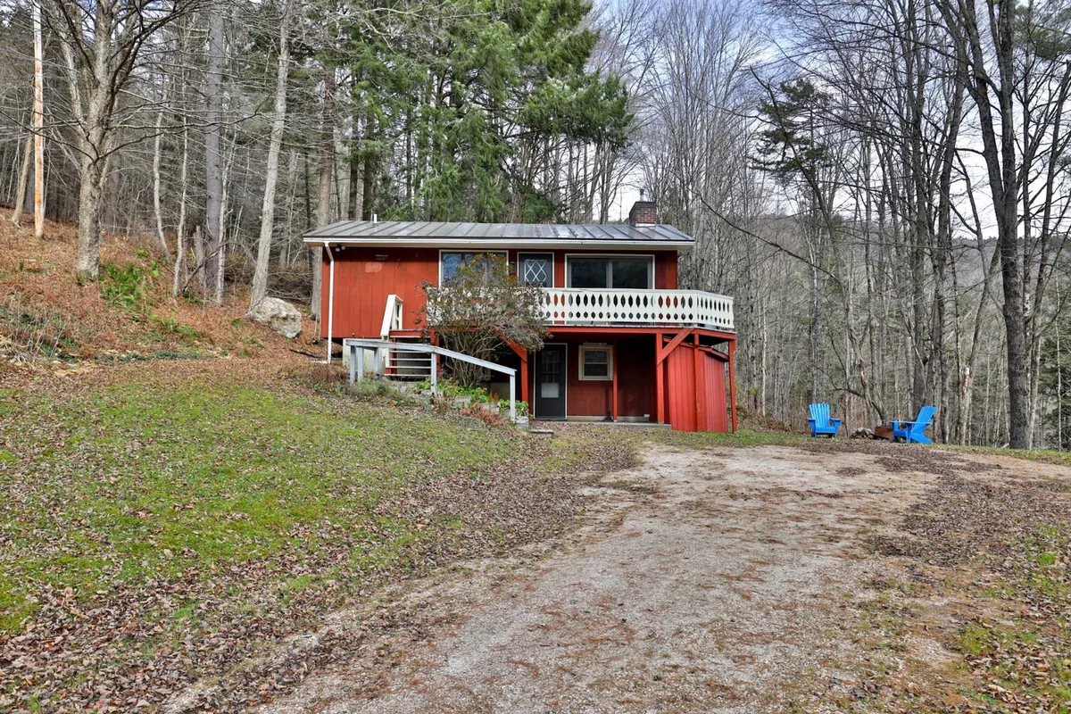 The classic red house sits beside a spacious deck furnished with blue chairs, surrounded by lush trees. The deck is set up for gathering, and the fire pit is nearby, illustrating the seamless transition from indoor comfort to outdoor enjoyment. Evening light creates a cozy, inviting mood.