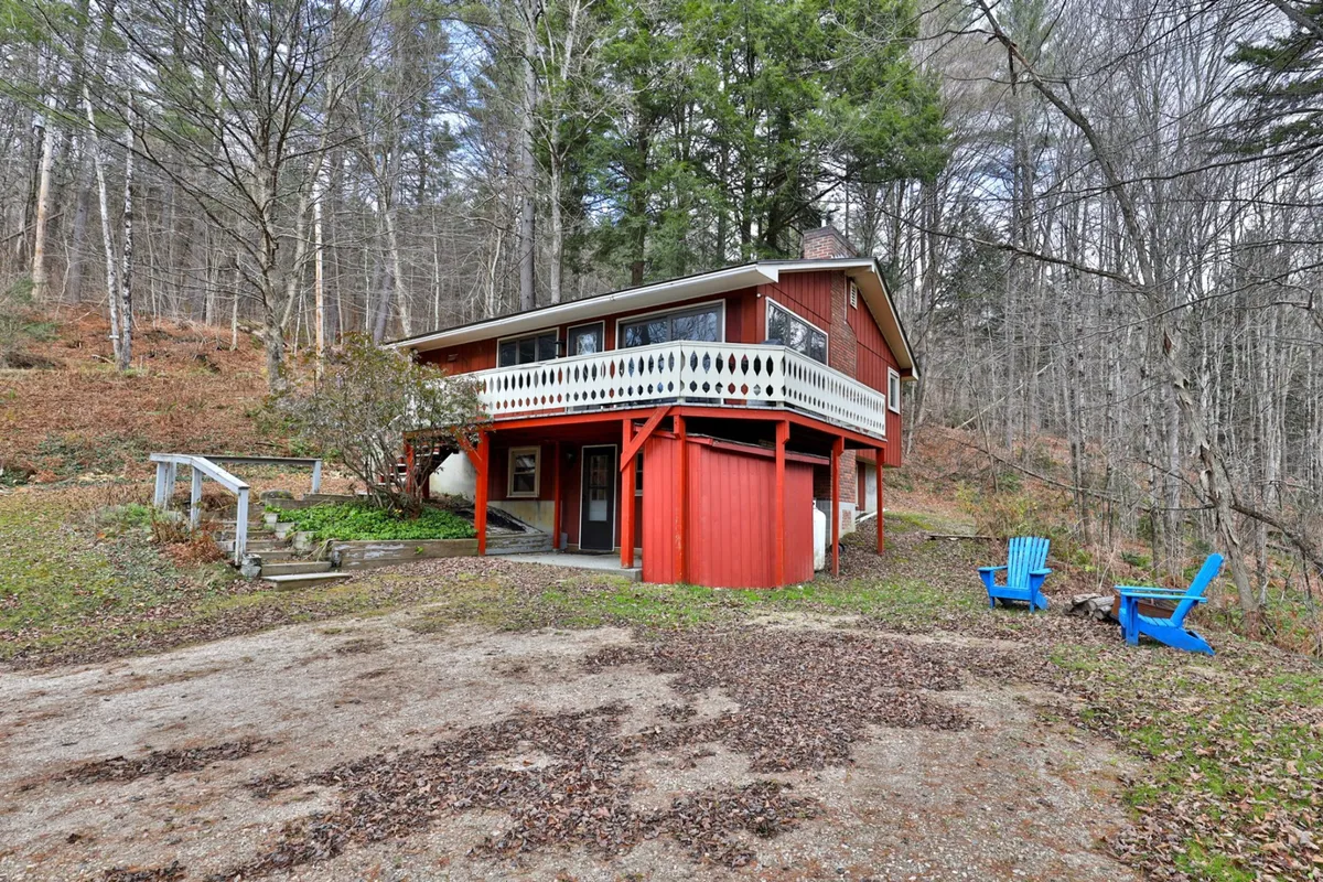 A vibrant red house nestled among snow-dusted trees, featuring a spacious balcony with blue chairs and a view of the surrounding winter landscape. The outdoor fire pit area is visible, inviting relaxation after a day in the snow.