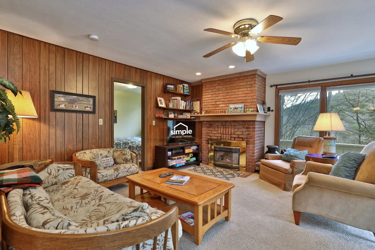 The image showcases a cozy living room with a crackling fireplace as the centerpiece, surrounded by a well-stocked bookshelf and a large window that frames peaceful mountain views. Comfortable seating invites guests to relax, read, or socialize while natural light fills the space, creating a warm and inviting atmosphere.