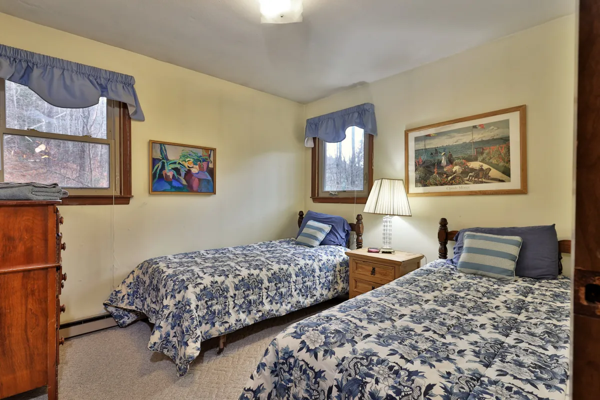 A bright bedroom with twin beds covered in cheerful floral bedding, warm lamps, artwork decorating the walls, and a window streaming in natural light. The room exudes a relaxed, inviting vibe suitable for groups sharing accommodations.