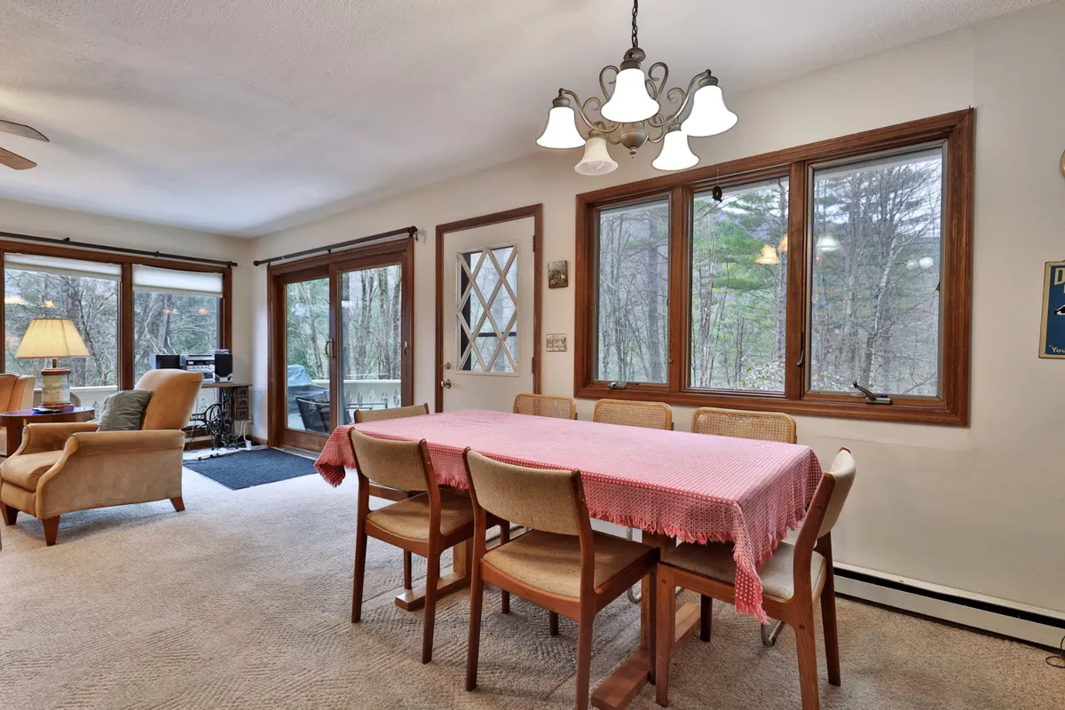 A bright and cozy dining area with a table, windows offering natural light and countryside views, a chandelier, a sofa, and a lamp. The setup invites both gatherings and quiet moments, with the outdoors visible through the windows.