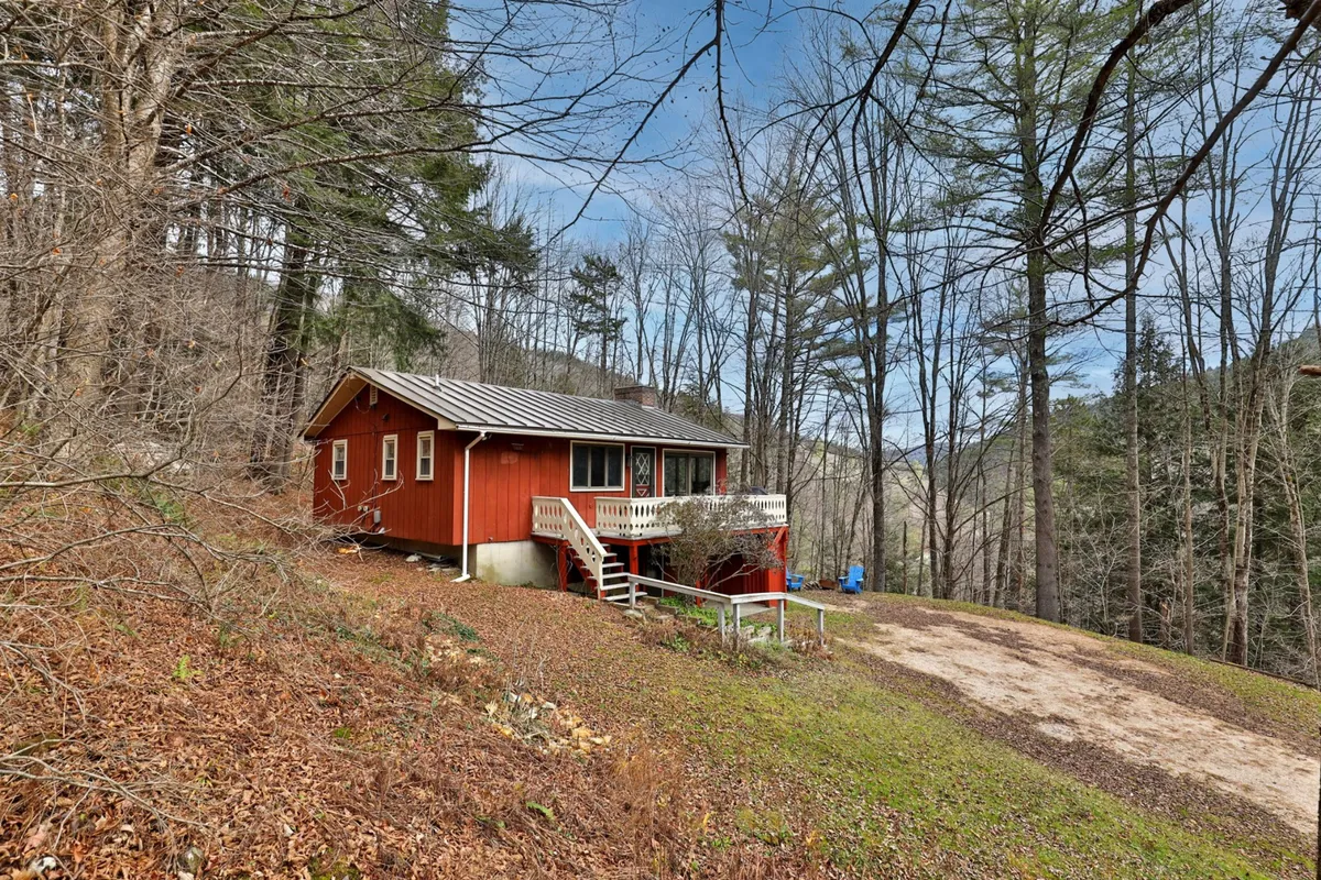 An inviting exterior view of a red cabin surrounded by lush green trees, with a welcoming front porch offering a peaceful place to relax. The image captures the connection between the property and its natural Vermont setting, with hints of nearby woods perfect for hiking and nature exploration.