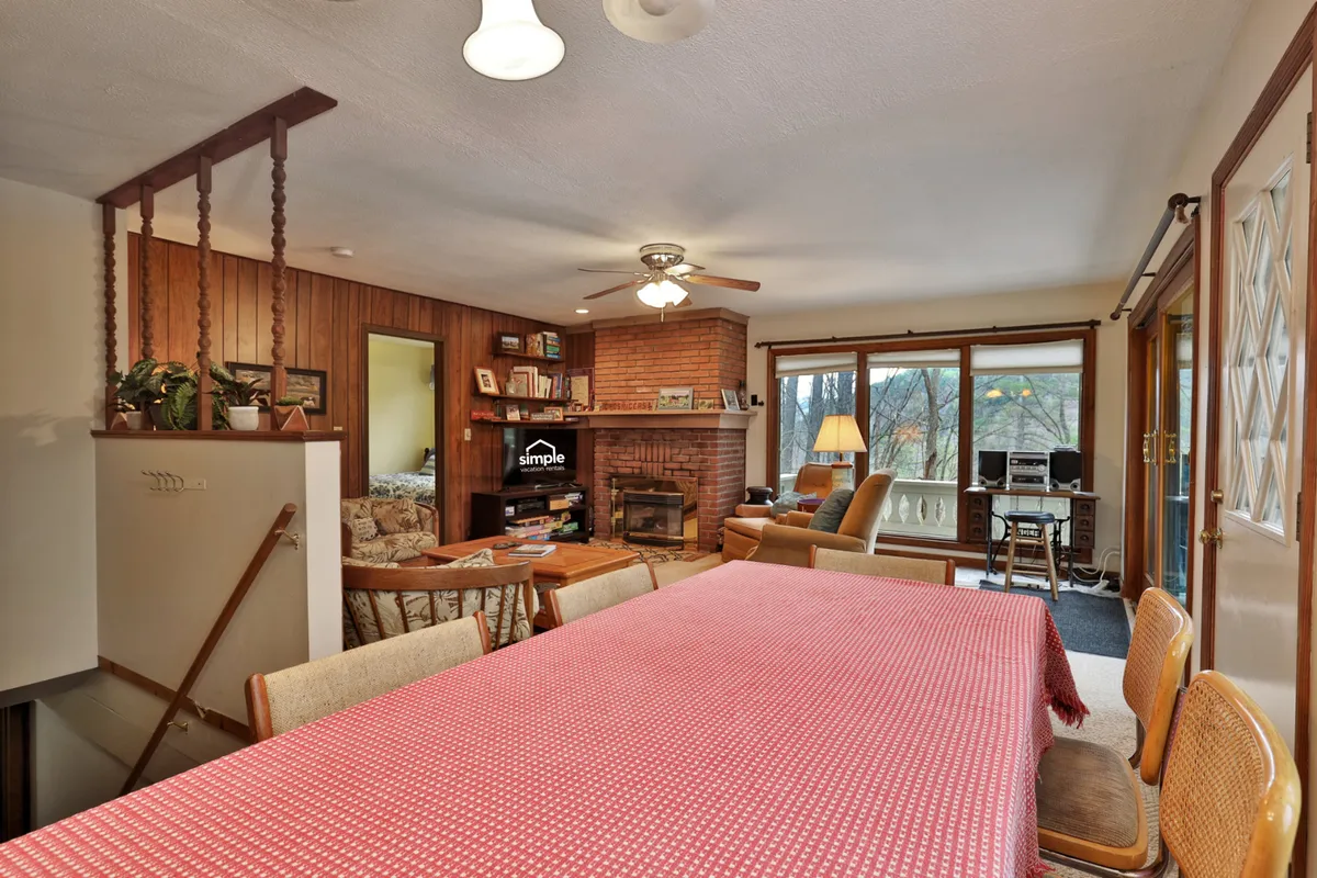 A spacious living and dining area featuring a large dining table with a red checkered tablecloth, a prominent fireplace, comfortable seating, and windows that offer views of the outdoors. The room is warm and welcoming, ideal for hosting family gatherings, sharing meals, or relaxing by the fire.