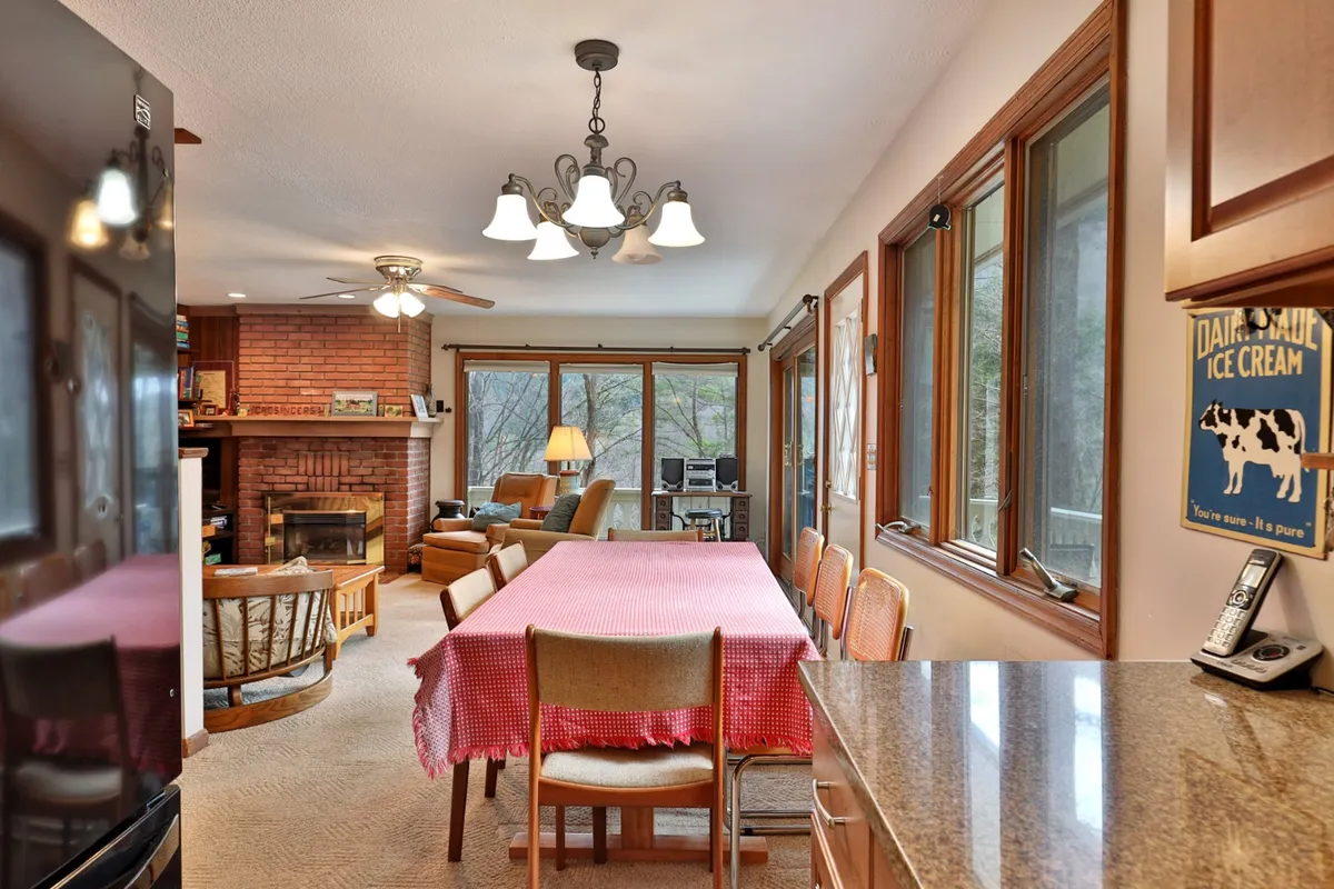 A large dining table draped in a checkered tablecloth sits beside a brick fireplace, surrounded by cozy seating and bathed in natural light from wide windows. The space feels designed for lively family meals, game nights, and heartfelt conversations.