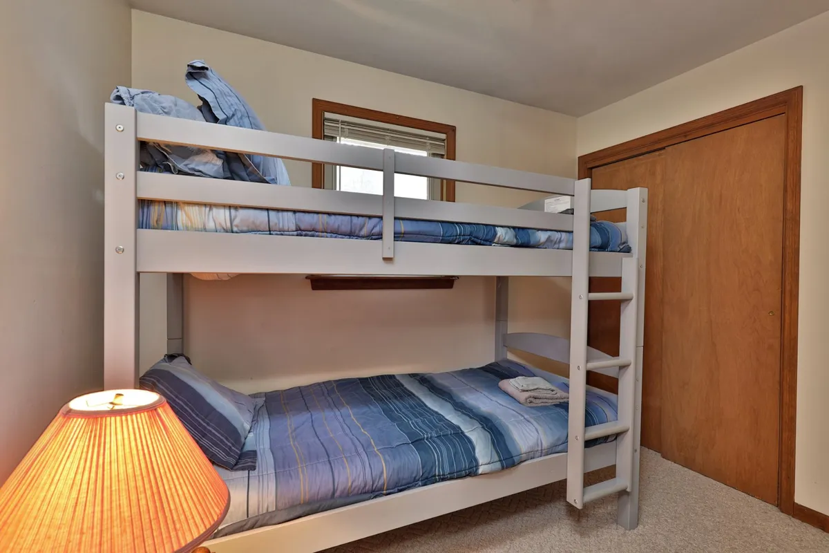 A playful bunk room with stacked bunk beds topped with cozy bedding, a lamp for nighttime reading, and a window letting in soft daylight. The space is inviting, especially appealing for families or groups with children.