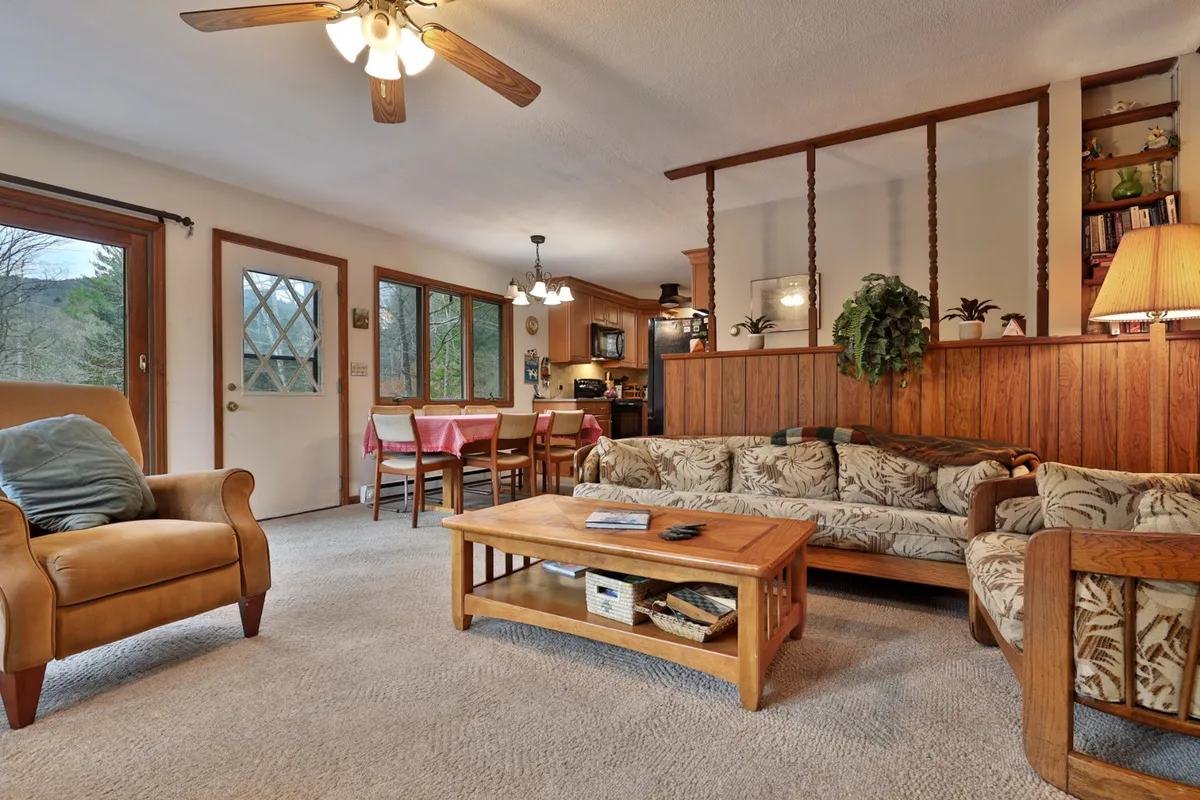 A warm and welcoming living and dining area featuring a sofa with a floral pattern, a wooden coffee table, and a dining area with red chairs. An open doorway visually connects the interior to the outdoors, hinting at the relaxing transition between indoor comfort and the surrounding countryside.